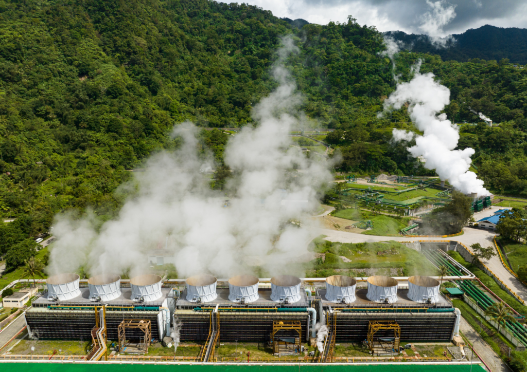 緑豊かな山々に囲まれた地熱発電所の航空写真。手前に冷却塔が複数並び、白い蒸気を上げている。奥には発電施設と配管が見える。周囲は緑の木々に覆われ、空には白い雲が点在している。